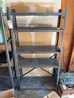 Front view of one metal shelving unit showing five shelves with some dust and dirt, placed against a garage wall.