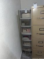 Two stackable plastic drawer units stacked against basement wall next to a beige filing cabinet. Each unit contains three translucent drawers with small assorted boxes inside.