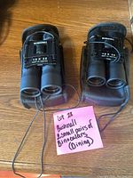 Two small Bushnell binoculars with cases placed side by side on a wooden surface with a pink note indicating lot information.