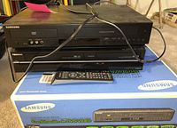 Photo showing Samsung DVD player on top of Toshiba DVD/VHS player with remote control on Samsung box.