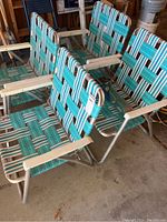 Four matching outdoor folding chairs with woven plastic webbing in turquoise, green, and white colors and aluminum frame with white armrests, folded in garage space.