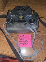 Front view of Bushnell binoculars with yellow branding, black body, carrying case underneath, and two neck straps.
