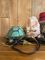 Photo showing green turtle-shaped lamp with cord and pink Himalayan salt lamp on wooden base on a wooden surface against wood paneling.