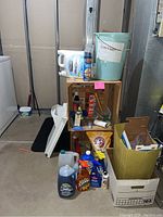 Full view of all cleaning and laundry items arranged on and around wooden crates including jugs, spray bottles, bucket, and cardboard box.