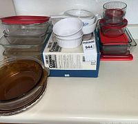 Kitchen counter with assorted glass bakeware including Corningware, Rubbermaid plastic lids, white ceramic ramekins, glass pie pans. All items stacked for displaying.