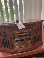 Front view of wooden multimedia speaker/player unit showing radio tuner, cassette player, and dual side speaker grills with intricate design.
