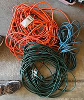 Three coiled extension cords in orange, blue, and green on a carpeted floor near a sneaker