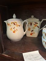 Photo showing cream pitcher with lid and sugar bowl with lid featuring floral orange and yellow leaf design on a white ceramic base inside wooden shelf.