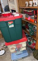 Large green storage bin with red lid stacked on top of clear and blue storage bins adjacent to shelves with Christmas wrapping paper and tins.