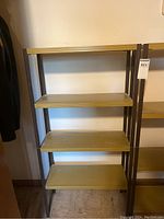 Photo showing one metal shelving unit with four yellowish-beige metal shelves and dark frame, standing against wall.