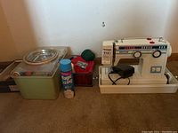 Wide shot showing the sewing machine, foot pedal, thread, and multiple containers on the floor against a white wall