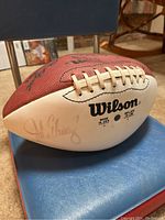 Wilson football with John Elway signature on white leather panel, shown on blue-red padded portable bleacher seat.