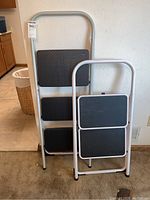 Front view of two stepladders standing next to each other, one taller with three steps and one shorter with two steps.