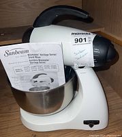Sunbeam Mixmaster Legacy Edition stand mixer with stainless steel bowl and user manual inside the bowl, placed in a wooden cabinet.