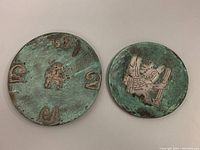 Two round wall plates showing oxidized copper surfaces with sterling silver inlays of indigenous Peruvian designs. One plate has symbols and a small figure, the other a winged figure with a staff.