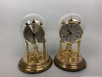 Two 9-inch vintage German glass domed mantle clocks under glass domes side by side on a white surface.