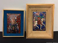 Two framed copper enamel religious plaques, one larger with beige frame and one smaller with gold frame, both showing Christian iconography with vibrant colors, set against blue velvet backing.
