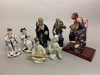 Full lot of nine Asian-style figurines including Mudmen, porcelain Emperor/Empress, musicians and kimono dancer.