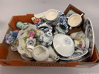 Wide shot of box containing miscellanea including porcelain teacups, saucers, small vases, and decorative pieces piled inside an orange box.