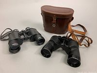 Photo showing two pairs of binoculars and one brown leather carrying case with strap and snap closure. One binocular pair is the Carl Wetzler item described.