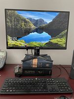 Black Acer widescreen LCD monitor powered on, showing a nature wallpaper. Setup includes black wired keyboard in front and cables connected at the back.