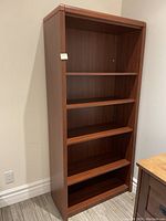 Wooden five-shelf bookcase with a reddish-brown finish, shown standing upright in a room corner, highlighting its size and shelf configuration.