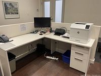 L shaped white modular desk and 3 drawer cabinet in office setting with monitor, printer, phone and office supplies on desk surface.
