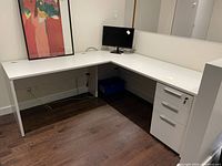White L shaped modular desk with a corner design and workspace on either side. The desk has a drawer unit on wheels underneath the right side, and is placed against white walls with dark hardwood flooring.