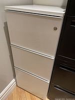 White 3-drawer filing cabinet in corner beside black furniture, showing lock cylinder on top drawer and clean laminate surfaces with minor signs of use.