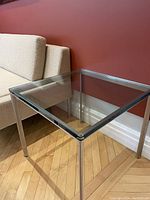 Square glass side table shown near beige upholstered chair and red wall on hardwood flooring. Clear view of thick solid glass top and brushed metal legs visible.