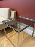Square glass side table next to a beige couch, showing the metal legs and wooden herringbone floor.