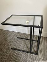 Front angled view of rectangular side table with glass top and black metal frame on carpeted floor next to white wall.