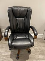 Front view of black faux leather office chair with padded back and seat, wood arms and caster base