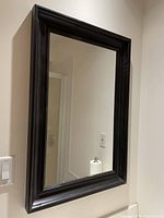 Photo of a rectangular wall mirror with a black wood frame hung on a light colored wall, showing the reflective glass and part of the frame's edge details.