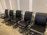 Five of six black leather office chairs arranged in a row showing seats, backs, bases and armrests.