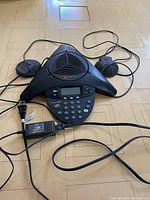 Polycom boardroom phone showing the central control panel with keypad, display, and two satellite microphones with cables, including power adapter.