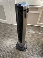 Full view of Seville Classic Tower Fan standing on carpeted floor, showing black grille and light gray body with attached remote control.