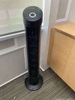 Front side view of black Seville Classics tower fan with visible vertical grill and round base, placed near window and wooden cabinet.