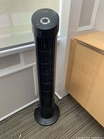 Full view of the black tower fan standing on the floor near a cabinet and window.