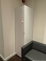 Closed white wardrobe closet next to gray armchair, showing its tall, narrow structure.