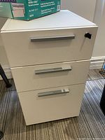 Front view of white laminate drawer unit with three drawers and metal handles, located on carpeted floor.