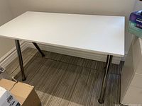 Full view of white rectangular laminate table with metal legs on a striped carpet floor next to wall and boxes.