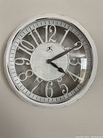 Front view of a white round wall clock showing stylized large white numbers on a gray background and black hands under clear glass