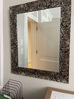 Square wall-mounted mirror with black and pewter mosaic wood frame hung on a white wall, reflecting part of the room.