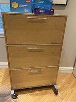 Front view of a wooden filing cabinet with three drawers and metal handles on caster wheels. Top holds boxes of file folders.