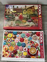 Photo showing two closed jigsaw puzzles boxes stacked - top shows cupcakes puzzle cover, bottom shows old town village puzzle cover