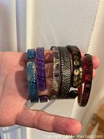 Photo showing 6 bracelets held in hand, various colors and materials including blue crackled resin and red marbled resin.
