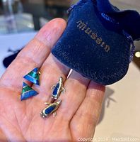 Two pairs of earrings held in hand next to a blue velvet Mussio pouch.