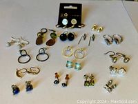 Front view of assortment of 19 pairs of costume jewelry post earrings in various styles including metallic, stone, pearl, and loop designs on flat surface.