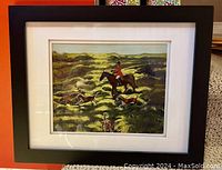 Framed print showing a foxhunt scene with a rider on horseback and dogs in a grassy field, black frame and white matting.